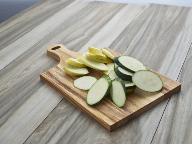 Slices of raw zucchini and summer squash in separate piles on a wooden paddle, close-up