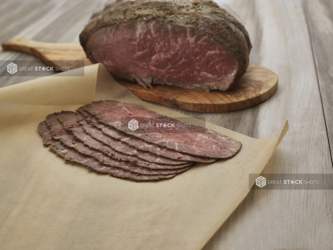 Thin slices of roast beef on brown parchment, close-up, remaining roast on wood paddle in background