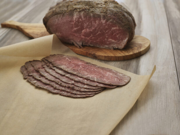 Thin slices of roast beef on brown parchment, close-up, remaining roast on wood paddle in background