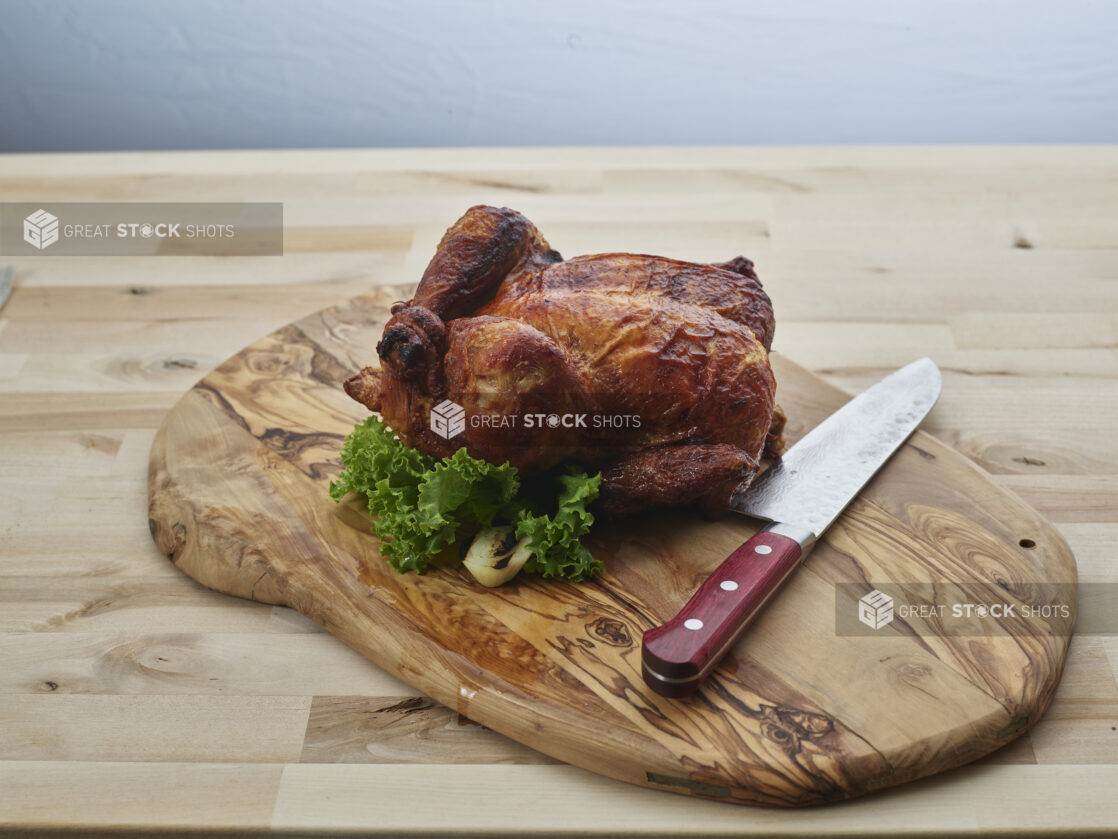 Small golden brown roasted chicken with carving knife on a natural rounded wood board