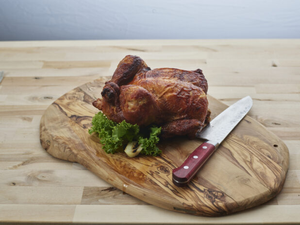 Small golden brown roasted chicken with carving knife on a natural rounded wood board