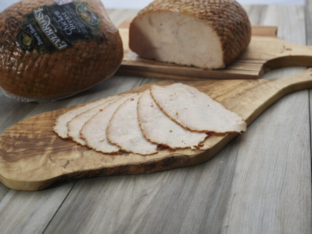 Thin slices of roast deli chicken arranged on a wooden paddle, whole and partial chicken breast roasts in background