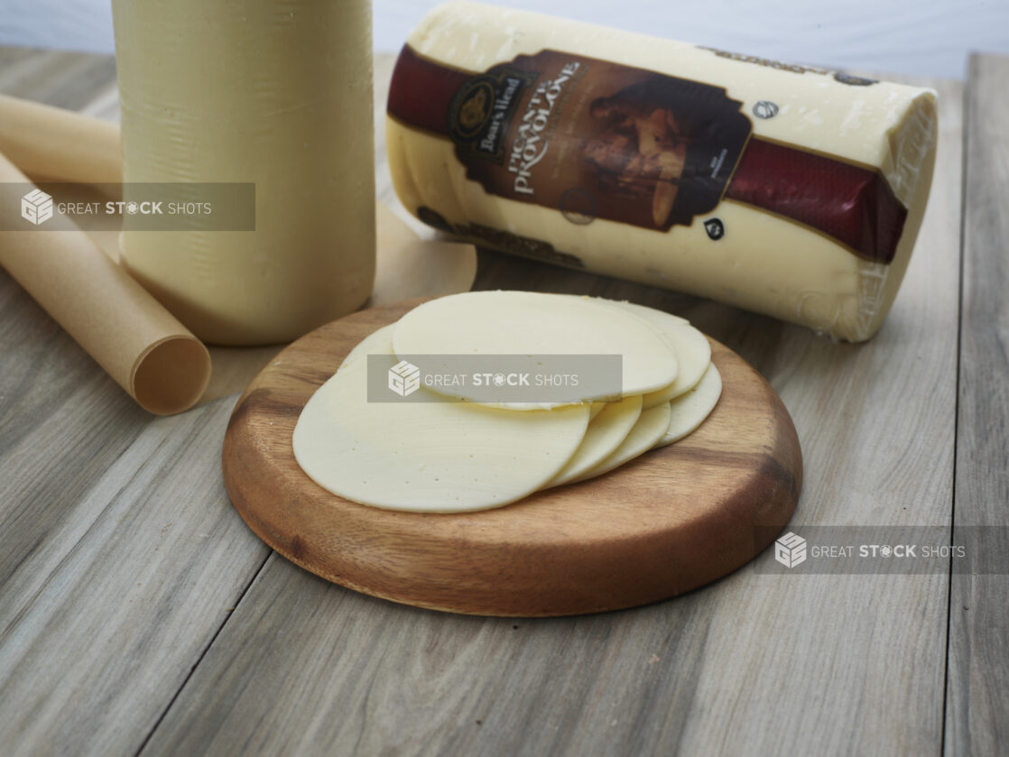 Sliced provolone cheese piled on a small round wood board, close-up, whole cheese in background