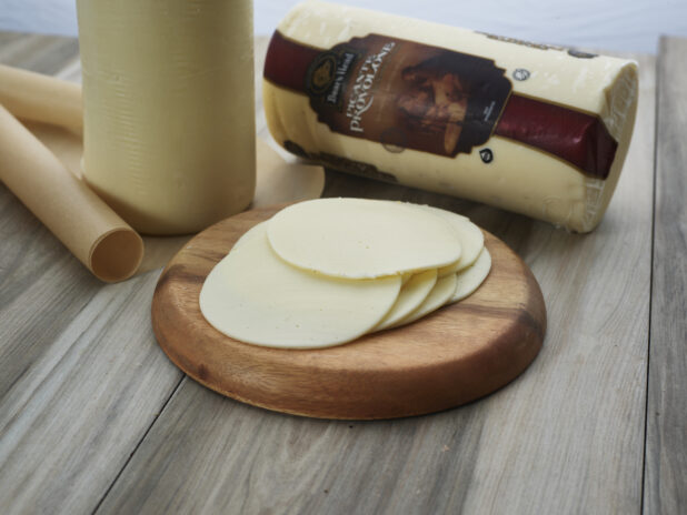 Sliced provolone cheese piled on a small round wood board, close-up, whole cheese in background