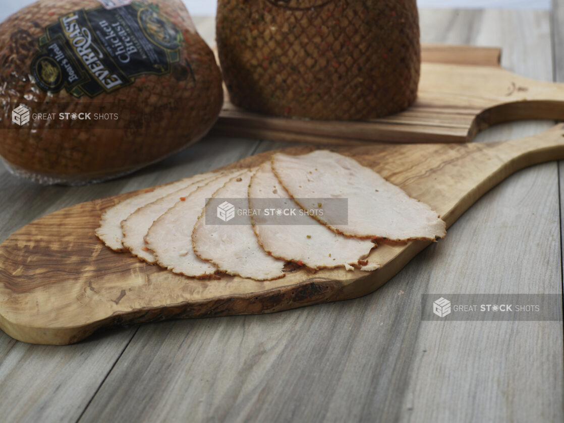 Thin slices of roast deli chicken arranged on a wooden paddle, whole wrapped chicken breast roasts in background