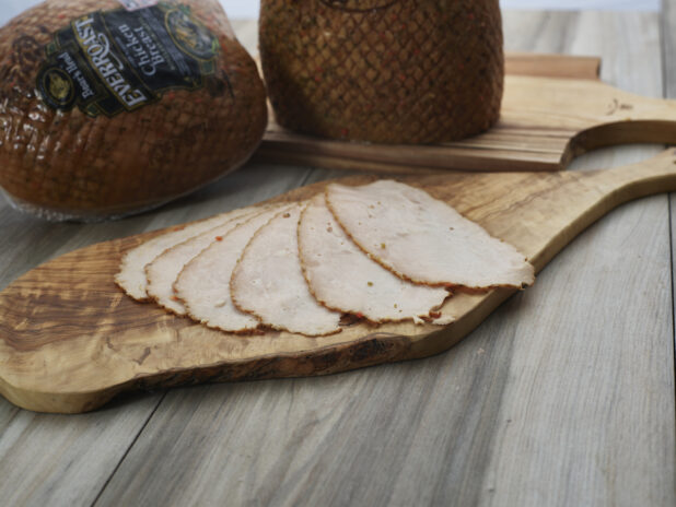 Thin slices of roast deli chicken arranged on a wooden paddle, whole wrapped chicken breast roasts in background