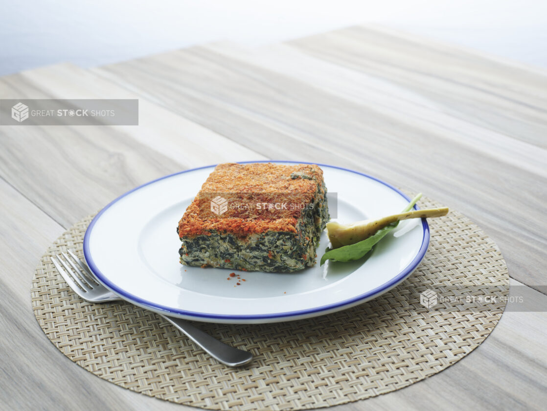 Square slice of spinach artichoke casserole with garnishes on a white and blue ceramic plate