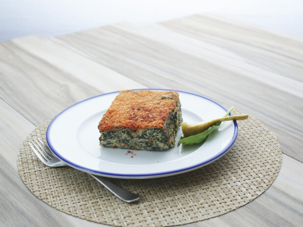 Square slice of spinach artichoke casserole with garnishes on a white and blue ceramic plate