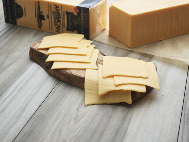 Sliced American cheese arranged on a natural wood board, whole block of cheese in background