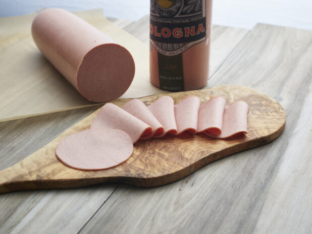 Bologna slices arranged on a wooden paddle, close-up, bologna sausage in background