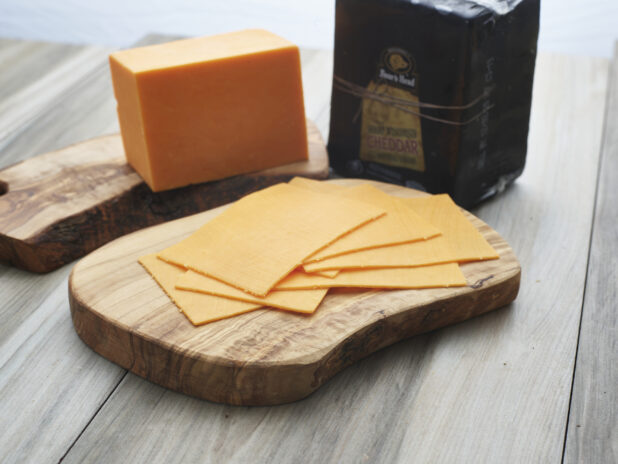 Slices of black wax cheddar cheese arranged on a wooden board, close-up, brick of cheese in background