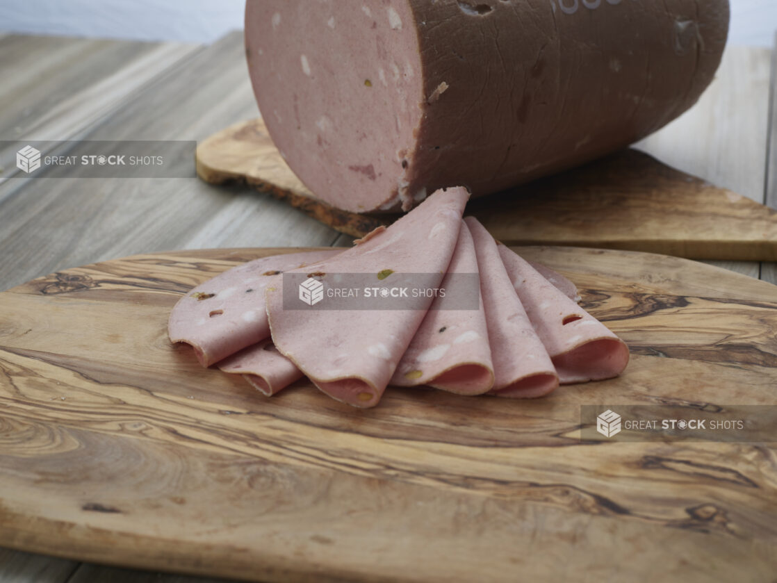 Thin slices of mortadella with pistachios arranged on a wood board, close-up, large piece in background