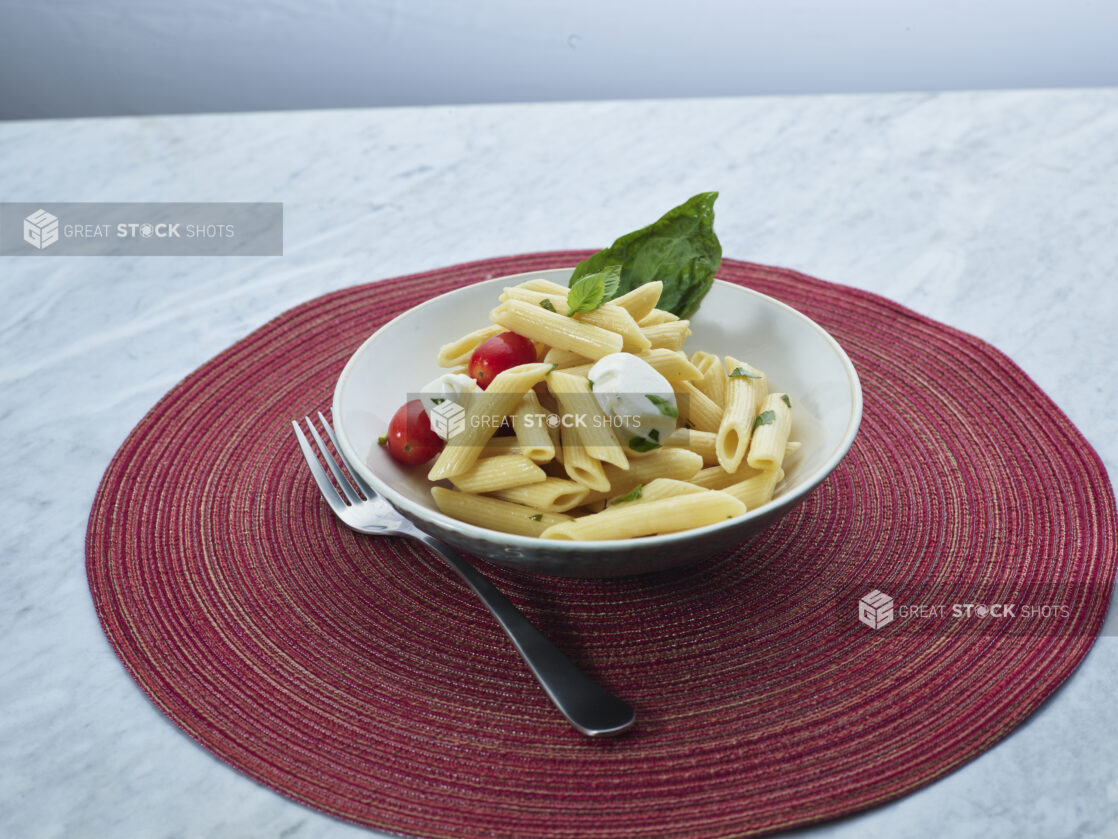 Bocconcini and tomato salad in a small white bowl on a round red woven placemat, white marble background