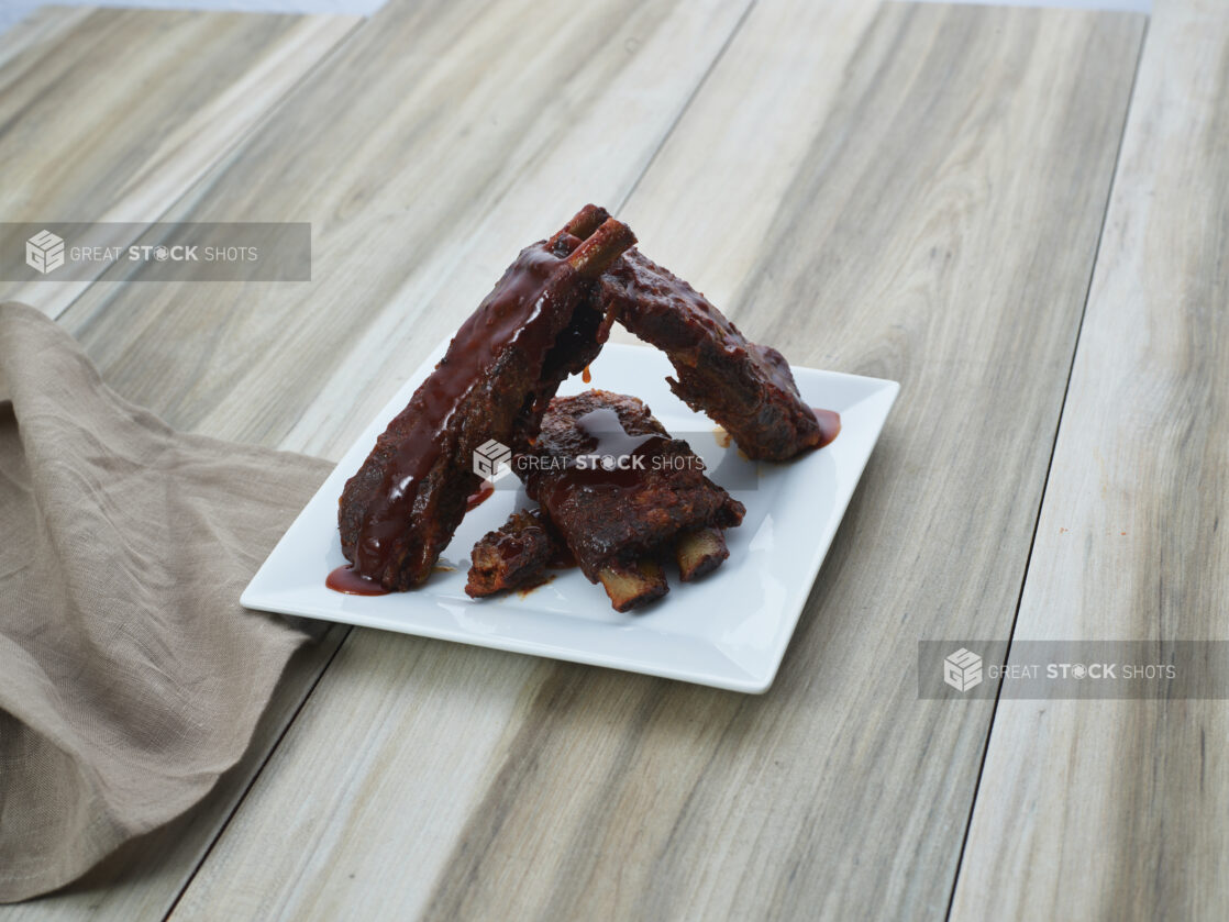 Sauced BBQ ribs arranged on a square white plate with cloth napkin, close-up