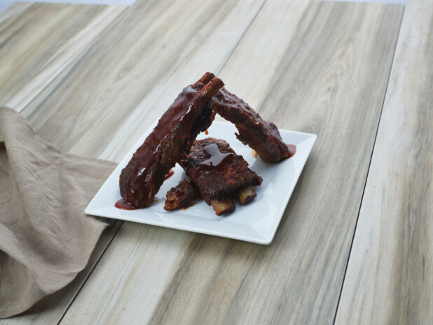 Sauced BBQ ribs arranged on a square white plate with cloth napkin, close-up