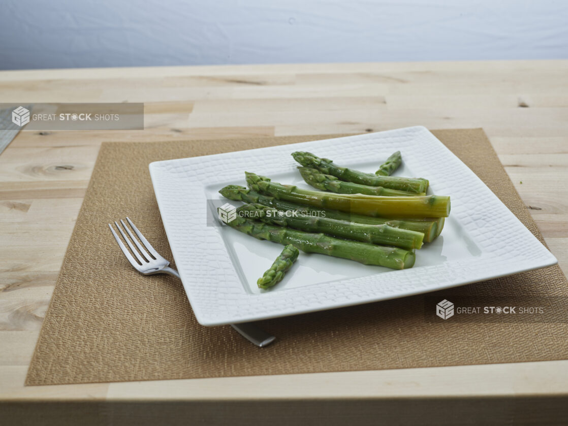 Steamed asparagus on a square white ceramic plate with a fork and grey woven placemat