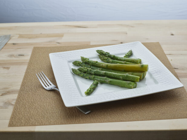 Steamed asparagus on a square white ceramic plate with a fork and grey woven placemat