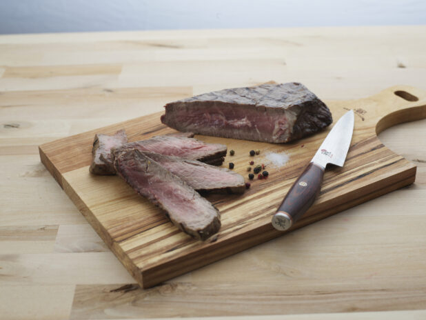 London broil steak partially sliced with seasonings and a Japanese knife on a wooden paddle