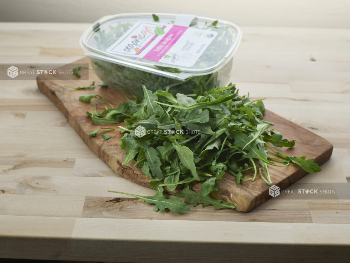 Clear plastic container of fresh arugula with some piled in front on a wood board, close-up