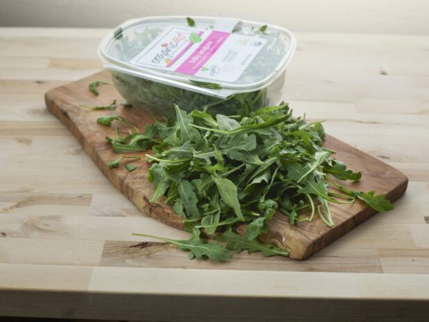 Clear plastic container of fresh arugula with some piled in front on a wood board, close-up