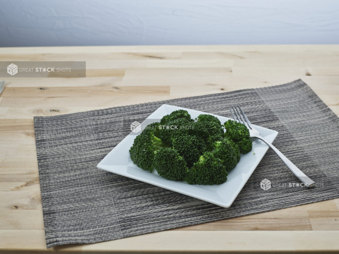 Steamed broccoli on a square white ceramic plate with a fork, grey woven placemat