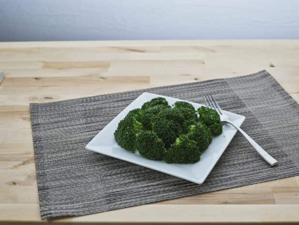 Steamed broccoli on a square white ceramic plate with a fork, grey woven placemat