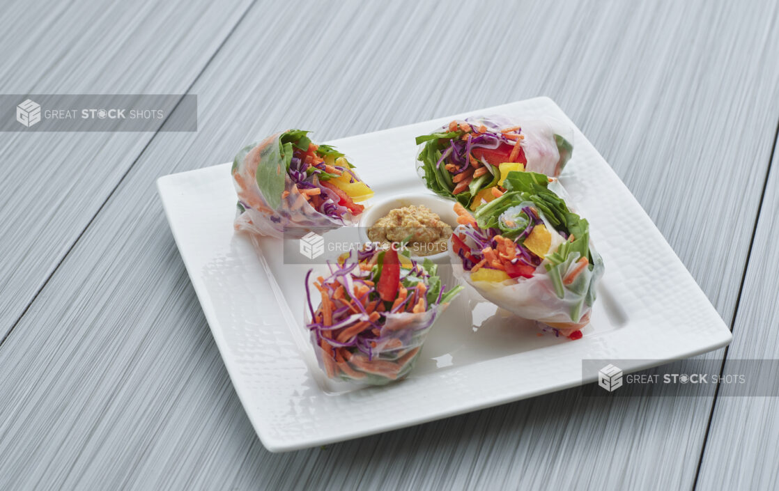 Four brightly-coloured fresh rice paper spring/summer rolls on a decorative square white plate, close-up