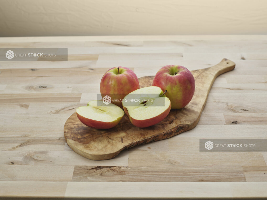 Three red and green apples, one sliced in half, on a wood cutting board