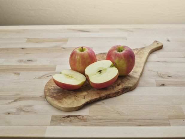 Three red and green apples, one sliced in half, on a wood cutting board