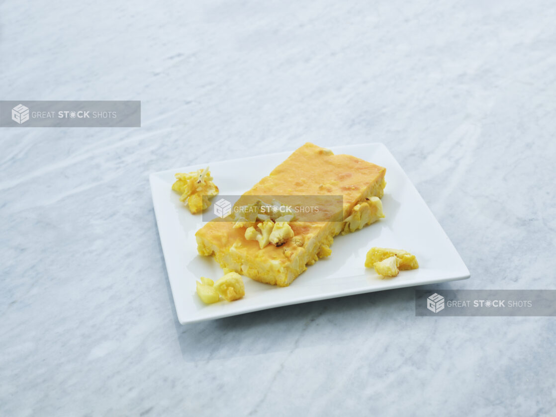 Rectangular slice of baked cauliflower macaroni and cheese on a square white plate, marble background