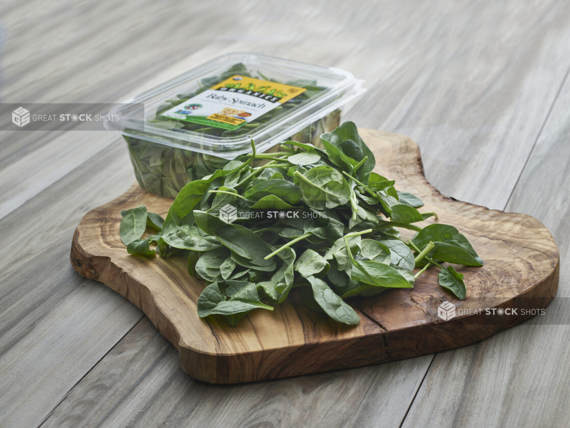 Fresh baby spinach piled on a natural wooden board, clear plastic grocery container in background