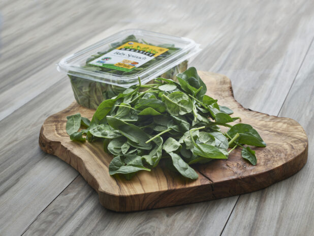 Fresh baby spinach piled on a natural wooden board, clear plastic grocery container in background