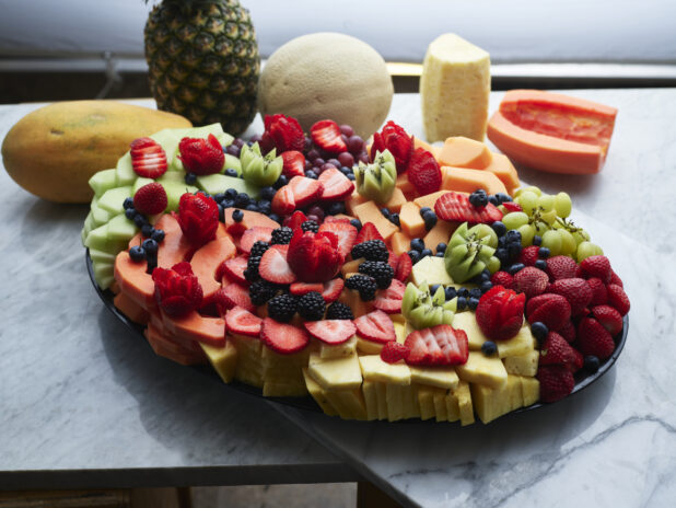 Luscious fruit arrrangement on black oval tray with various fruits behind, white marble background