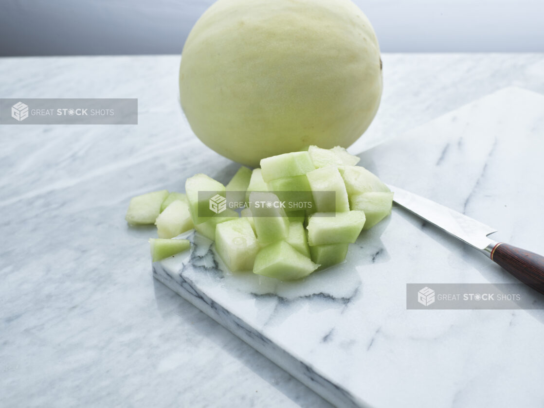 Chopped honeydew melon with a knife on a marble board with whole melon behind, white marble background