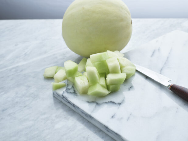 Chopped honeydew melon with a knife on a marble board with whole melon behind, white marble background