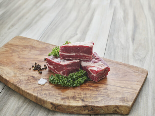 Three uncooked short ribs stacked on a wood cutting board with seasonings