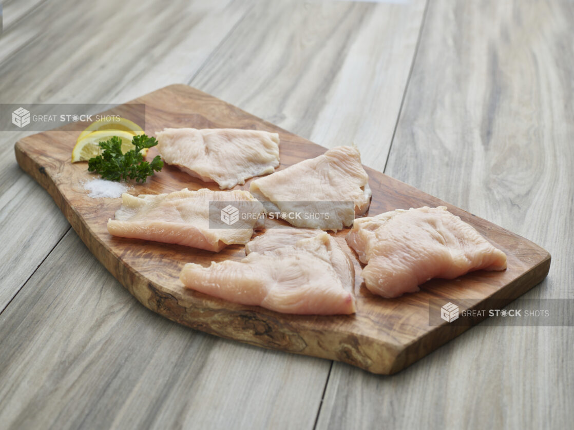 Five thinly sliced chicken cutlets with seasonings on a natural wood board, close-up