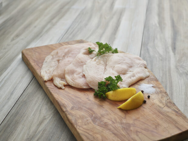 Thinly sliced raw chicken cutlets with seasonings on a natural wood board, close-up