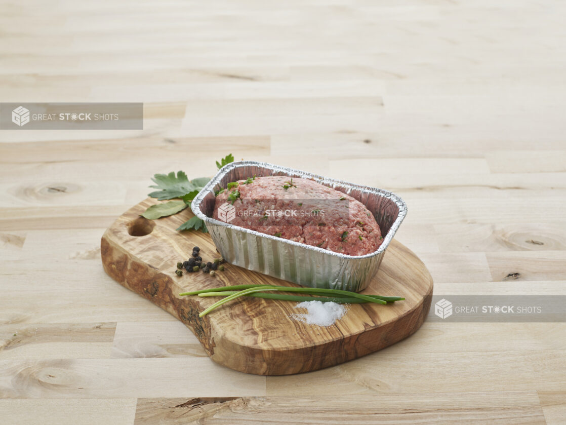 Ready-to-bake meatloaf, uncooked, in a foil baking pan with seasonings on a wood board