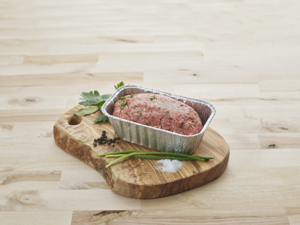 Ready-to-bake meatloaf, uncooked, in a foil baking pan with seasonings on a wood board