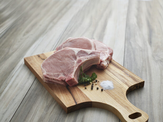 Two thick center cut pork chops with seasonings on a wooden paddle