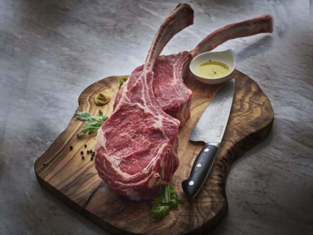 Two raw beef tomahawk steaks on a wooden cutting board with kitchen knife and seasonings, close-up