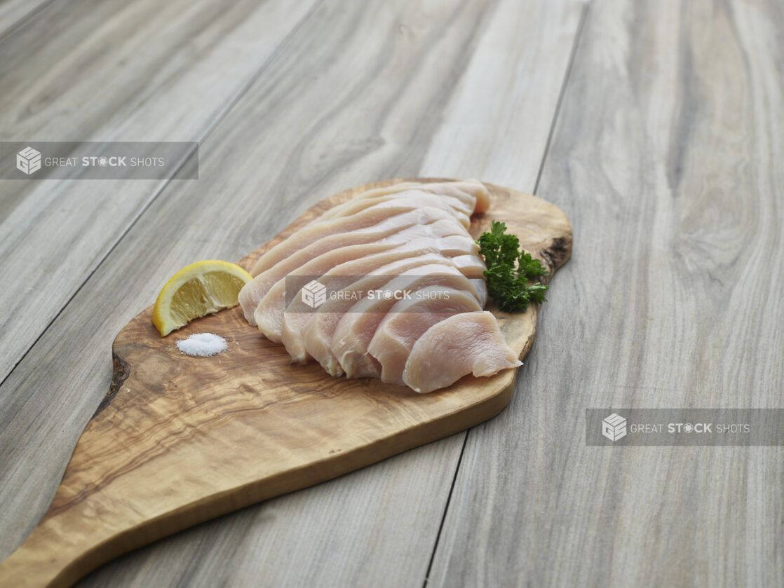 Sliced raw chicken breast fillet arranged on a natural wood board with seasonings