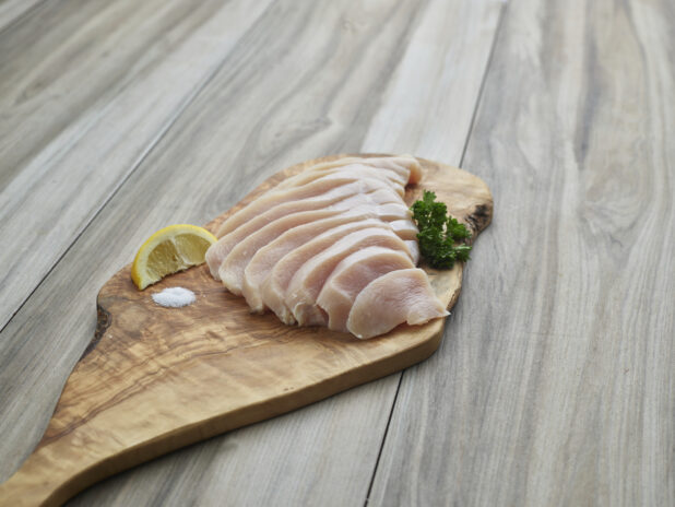 Sliced raw chicken breast fillet arranged on a natural wood board with seasonings