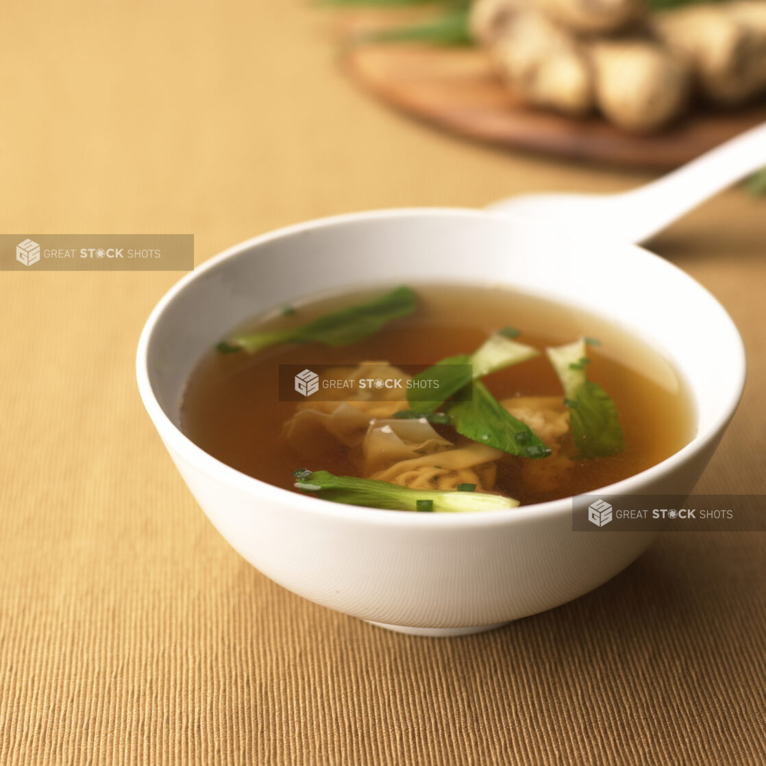 A White Ceramic Bowl of Wonton Soup with Baby Bok Choy on a Light Brown Tablecloth Surface