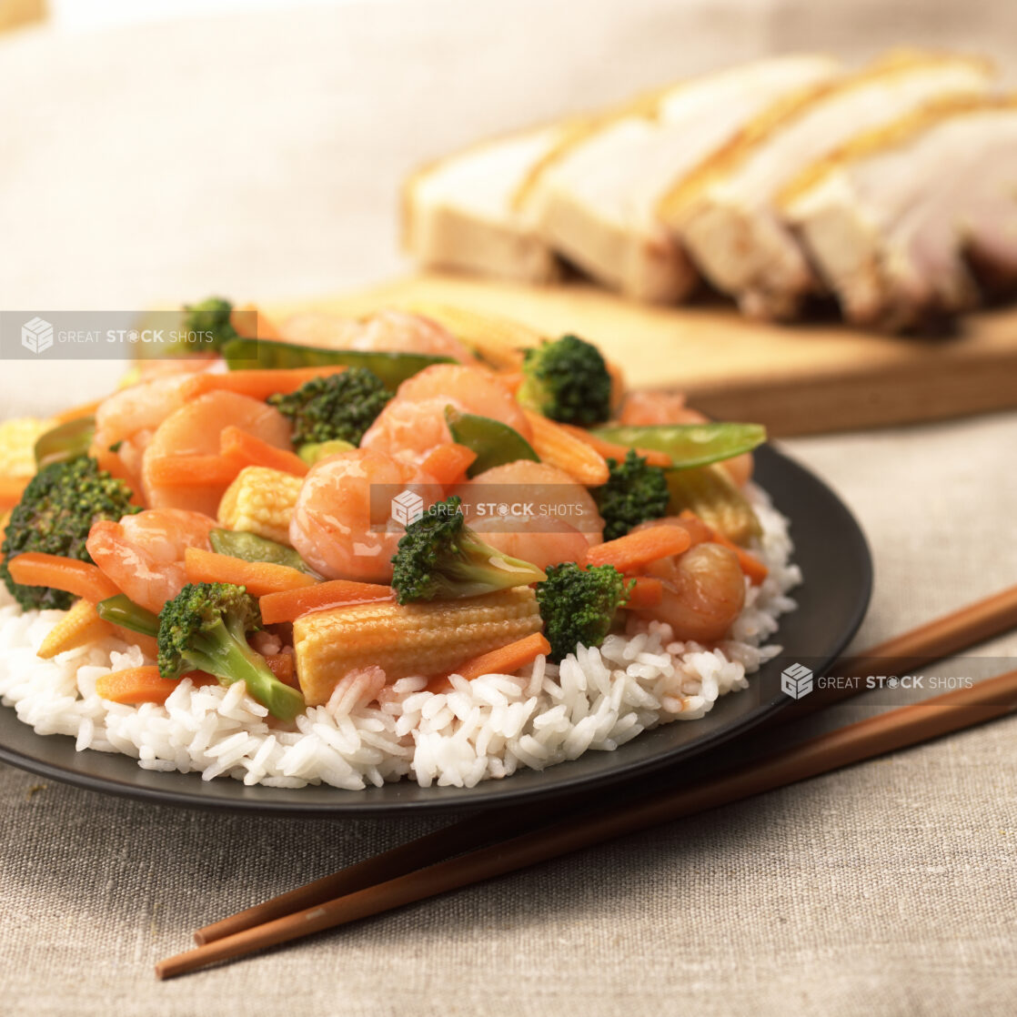 Close Up of a Dish of Shrimp and Vegetable Stir Fry Over White Rice on a Black Ceramic Plate with Crispy Pork Belly in the Background