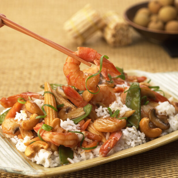 Close Up of a Dish of Shrimp, Vegetable and Cashew Stir Fry Over Rice With a Pair of Chopsticks Picking Up a Shrimp - Variation