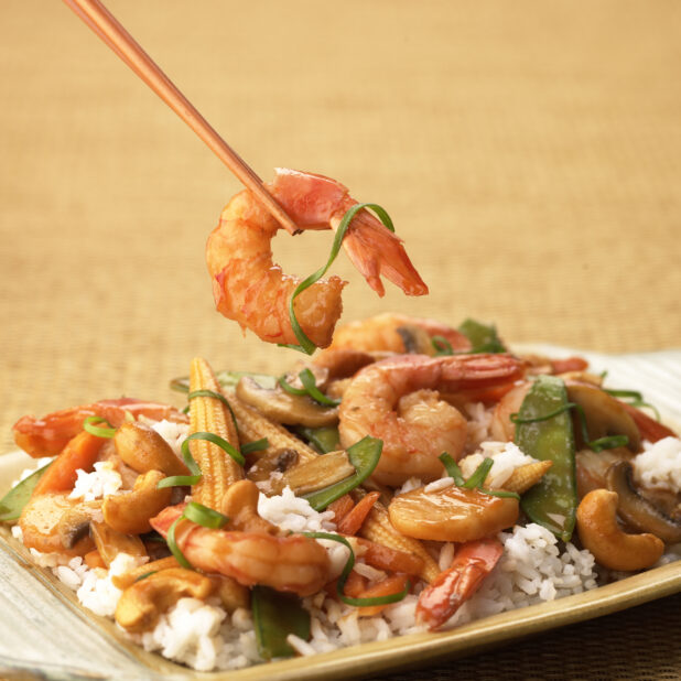 Close Up of a Dish of Shrimp, Vegetable and Cashew Stir Fry Over Rice With a Pair of Chopsticks Picking Up a Shrimp