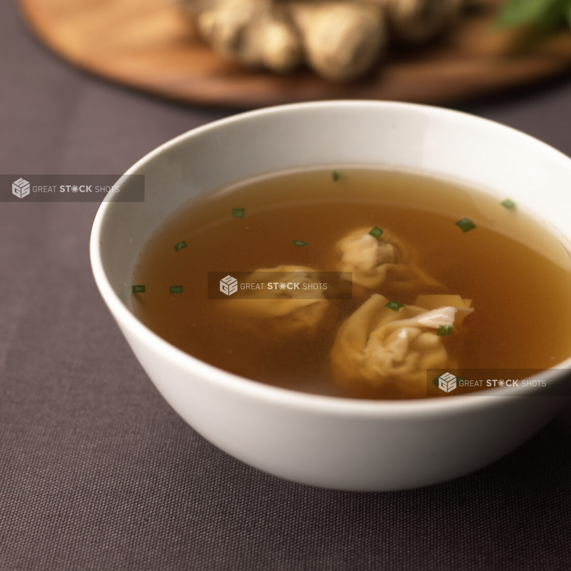 A White Ceramic Bowl of Wonton Soup with Chopped Chive Garnish on a Dark Brown Tablecloth Surface