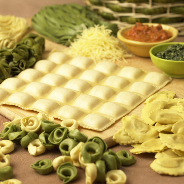 Assorted Fresh Pastas – Ravioli, Spinach Tortellini, Mezzalune and Linguine – on a Wooden Cutting Board in a Kitchen Setting